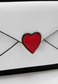 Biała torebka kopertówka z faux skóry z czarnymi wykończeniami, z centralnym akcentem w postaci czerwonego serca ze skóry i przeszywaną klapą w stylu koperty.