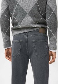 Jeans de mezclilla gris con un diseño ajustado, que presentan dos bolsillos traseros y un parche de cuero en la cinturilla, combinados con un suéter gris con patrón.
