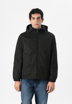 Svart huva jacka med ett texturerat geometriskt mönster, frontdragkedja och elastiska ärmslut. Parat med blå denimjeans.