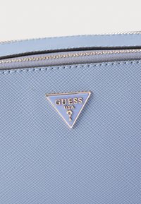 Φωτεινό γαλάζιο υλικό συνθετικής υφής με φερμουάρ και χρυσό τρίγωνο λογότυπο Guess. Τονίζεται από ένα λείο, αντίθετο μπλε φόντο.
