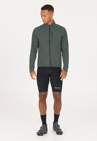 Letvægts, mørkegrøn cykeljakke med ståkrave, frontlynlås og en brystlomme; parret med sorte cykelshorts og sko.