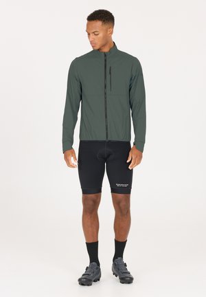 Leichte, dunkelgrüne Radjacke mit Stehkragen, Frontreißverschluss und Brusttasche; kombiniert mit schwarzen Fahrradhosen und Schuhen.