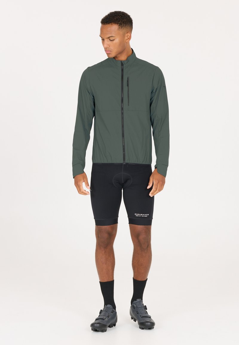 Letvægts, mørkegrøn cykeljakke med ståkrave, frontlynlås og en brystlomme; parret med sorte cykelshorts og sko.