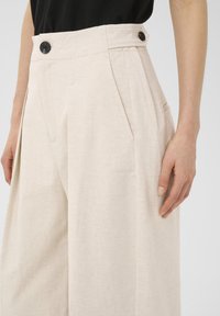 Beige wide-leg trousers made of textured fabric, featuring a high waist with two black buttons, side pockets, and pleats for a tailored look.