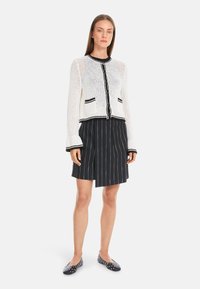 White knitted cardigan with black trim and buttons, paired with a black pinstriped skirt and polka-dot flats. Cropped design and loose fit.