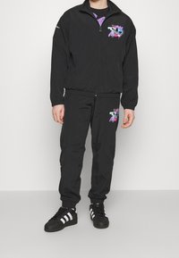 Svart träningsjacka och byxor i lätt material, med färgglada grafiska mönster på båda delarna. Skorna är svarta med vita ränder.