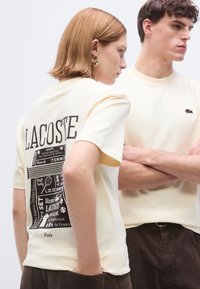 Κίτρινο βαμβακερό μπλουζάκι με μαύρο γραφικό τύπωμα στην πλάτη, που απεικονίζει στοιχεία του τένις και το κείμενο "LACOSTE". Το μοντέλο φορεί το μπλουζάκι σε συνδυασμό με μπεζ πουκάμισο.