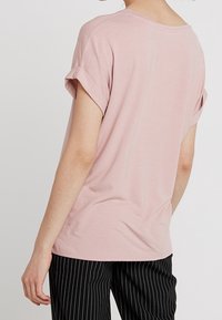 Person iført en lys pink kortærmet skjorte med opslåede manchetter og sorte stribede bukser, set bagfra mod en ensfarvet baggrund.