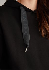 Sweat à capuche noir avec un large cordon noir présentant un texte subtil répété et un embout métallique, porté par une personne aux cheveux bruns.