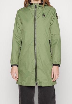 Kvinde iført en lang, lysegrøn hættetrøje med sorte lynlås- og lommedetaljer, kombineret med mørke bukser mod en ensfarvet baggrund.