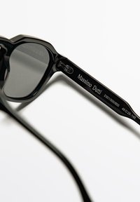 Schwarze Sonnenbrille mit geometrischem Rahmen und dunklen Gläsern. Mit graviertem Branding und Maßangaben am Bügel. Glatte, polierte Oberfläche.