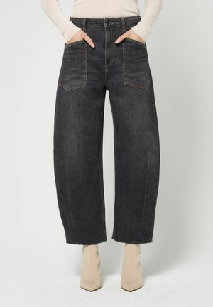 Person trägt schwarze Jeans mit hoher Taille und weitem Bein mit großen Vordertaschen, kombiniert mit beigefarbenen Absatzstiefeln und einem hellen langärmligen Oberteil.