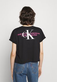 Persona con cabello rizado corto que lleva una camiseta corta negra de Calvin Klein y jeans de mezclilla azul, de espaldas a la cámara.