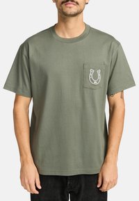 Camiseta de algodón verde oliva con cuello redondo, mangas cortas y un bolsillo frontal con un diseño de herradura blanca.