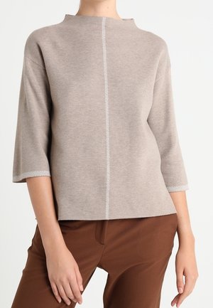 Femme portant un pull en maille beige à manches trois-quarts avec un bord blanc, associée à un pantalon marron sur un fond uni.