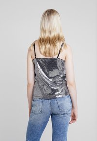 Femme blonde portant un haut sans manches en sequins argentés et un jean bleu, se tenant dos à l'appareil photo sur un fond uni.