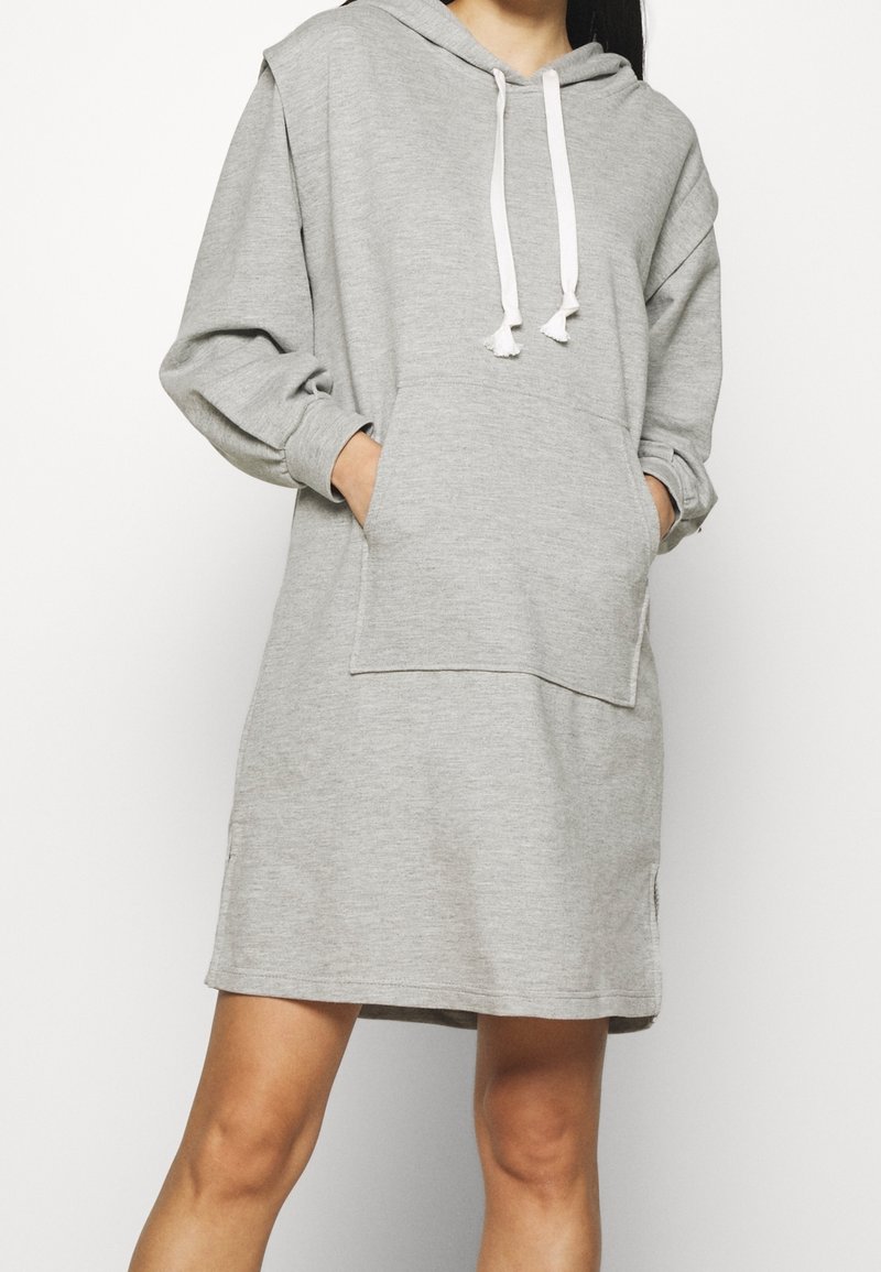 Femme portant une robe sweat à capuche ample de couleur gris clair avec poche avant et cordons blancs, les mains glissées dans la poche.