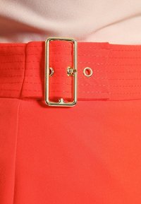 Červený textilní pásek se širokou obdélníkovou zlatou sponou, se švy a kovovým okem pro úpravu délky.