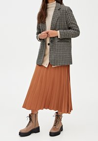 Blazer a scacchi in bianco e nero, dolcevita beige, gonna plissettata color ruggine e stivali marroni con lacci e suola spessa.