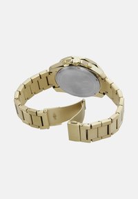 Guld metal armbåndsur med åben lås, der viser bagsiden af urkassen og to sideknapper på urkroppen.