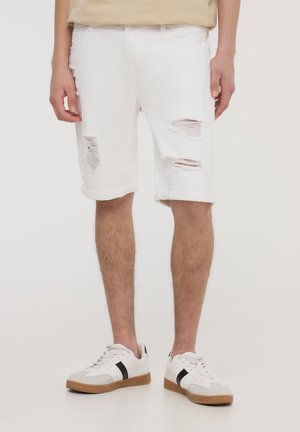 Shorts in denim bianco con dettagli strappati, orli arrotolati e una vestibilità rilassata, abbinati a sneakers bianche con una striscia nera.