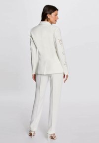 Completo bianco su misura con giacca aderente caratterizzata da ricami floreali sulle maniche e pantaloni dritti. Tessuto liscio e strutturato.