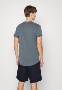 Tricou gri cu maneci scurte și margine rotunjită. Țesătura pare moale și ușoară. Asortat cu pantaloni scurți negri, evidențiind un aspect casual.
