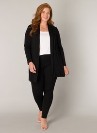 Zwarte lange cardigan over een witte top en zwarte leggings; gladde stof, open voorkant ontwerp, met lichte sandaaltjes.