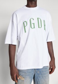 Vit bomulls t-shirt med fet grön "PGDR" text, korta ärmar, avslappnad passform och rundad halsringning. Bärs med slitna jeans.