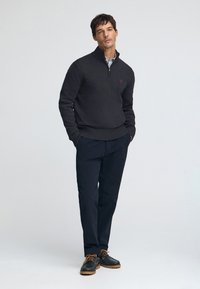 Dunkelgrauer Herrenstrickpullover mit Reißverschlusskragen, kombiniert mit marineblauen Hosen und dunklen Schuhen mit braunen Schnürsenkeln und strukturiertem Absatz. Rotes Logodetail.