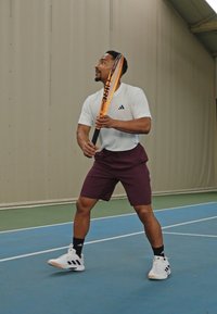 Joueur de tennis en chemise blanche à manches courtes, avec un short bordeaux et des chaussettes noires, tenant une raquette orange, positionné sur un court bleu.