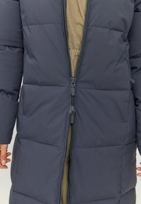 Doudoune bleu marine foncé, dotée d'une fermeture zippée devant, d'un design matelassé et d'une doublure intérieure douce couleur kaki. Tissu résistant avec sections matelassées.