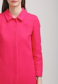 Kuuma roosa takki, jossa on tekstuuripinta, varustettu nappikauluksella ja pitkillä hihoilla. Suora leikkaus ja piilotettu etuosa.