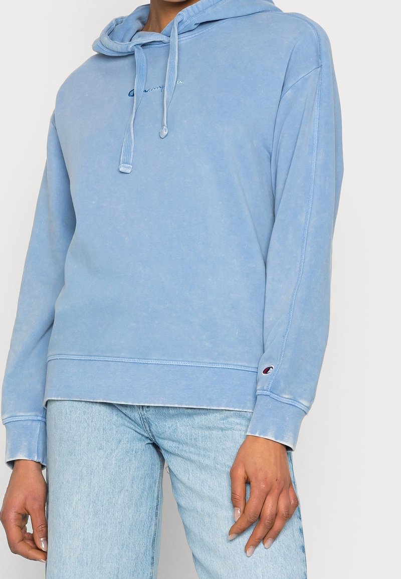 Personne portant un sweat à capuche Champion bleu clair avec cordons de serrage et un jean bleu clair, debout les mains détendues le long du corps.
