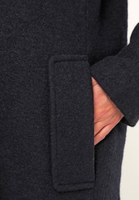 Gros plan sur un manteau en laine bleu marine montrant le tissu texturé, une poche latérale et une main reposant à l'intérieur de la poche. Aspect doux et chaud avec des lignes épurées.