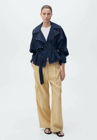 Jeansjasje met oversized revers en een gestrikte taille, gecombineerd met beige broek met wijde pijpen en cargozakken. Zwarte sandalen maken de outfit compleet.