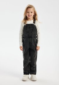Black denim overalls with adjustable straps, front pocket, and side buttons, paired with a long-sleeve white shirt and white sneakers.