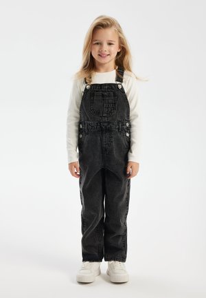 Pantalones de mezclilla negros con tirantes ajustables, bolsillo frontal y botones laterales, combinados con una camiseta blanca de manga larga y zapatillas blancas.