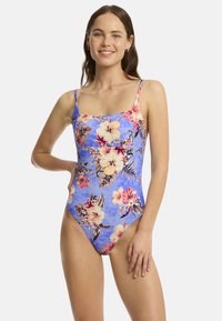 Une femme souriante pose dans un maillot de bain une pièce à fleurs avec un fond bleu, mettant en valeur des fleurs vibrantes et une expression détendue et joyeuse.
