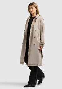 Naine seisab, kandes beeži värvi pika topeltrinnalise trentsi musta topi ja mustade laia säärega pükste ning mustade teravate saapade kohal.