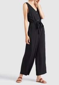 Black sleeveless jumpsuit with v-neck and buttons, tied waist, wide legs, made from smooth fabric; paired with brown slide sandals.