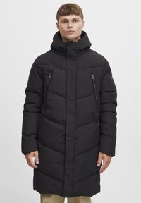 Geacă puffer neagră cu guler înalt, buzunare cu fermoar și o siluetă lungă. Are o textură matlasată și un design de culoare uniformă.