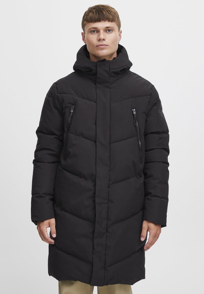 Geacă puffer neagră cu guler înalt, buzunare cu fermoar și o siluetă lungă. Are o textură matlasată și un design de culoare uniformă.