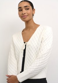 Cardigan tricoté blanc avec une texture en losanges, décolleté en V et fermeture à cordon. Manches et ourlet côtelés, en mélange de laine douce.