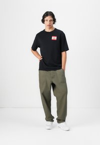 Sort bomulds T-shirt med rødt logo, parret med khaki vidde bukser. Modellen bærer hvide sneakers. Enkel, moderne design med afslappet pasform.