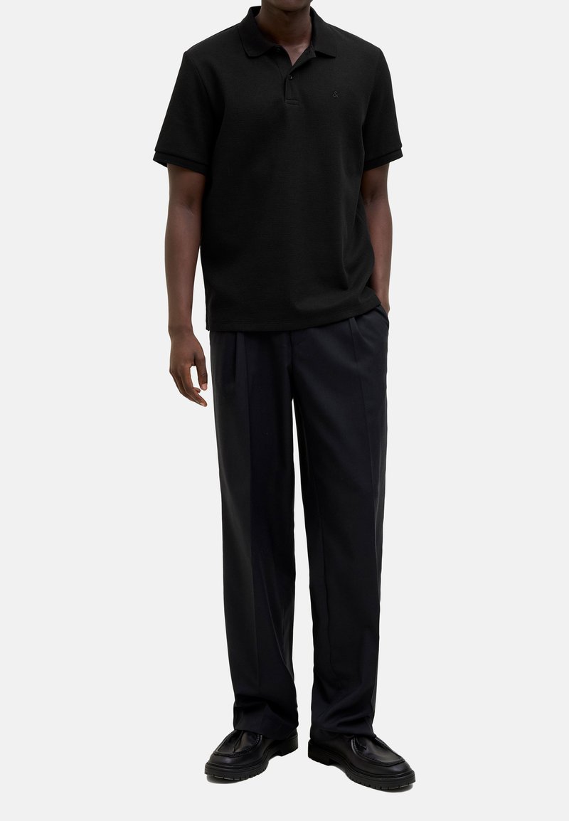 Polo shirt noir en tissu texturé, à manches courtes et col, associé à un pantalon noir ample et des chaussures noires.