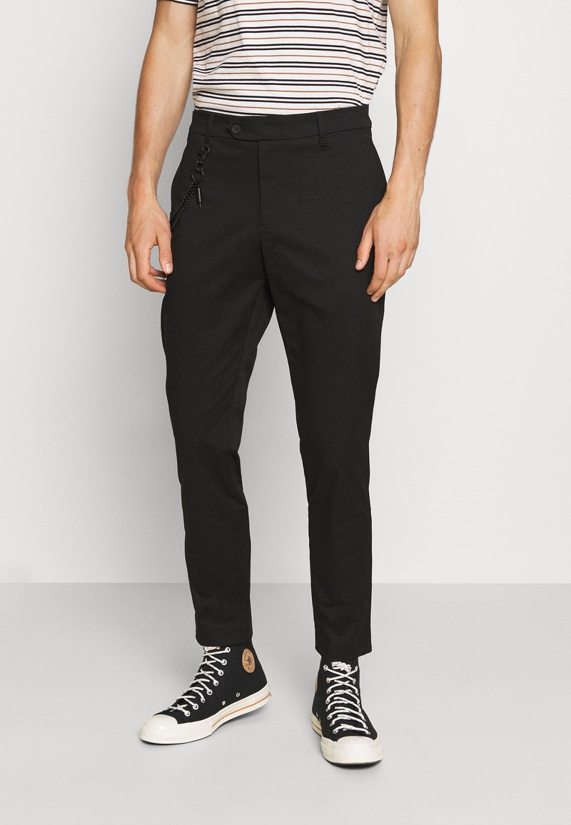 Homme portant un pantalon slim noir avec un accessoire chaîne, associé à des baskets montantes noires et une chemise rayée.
