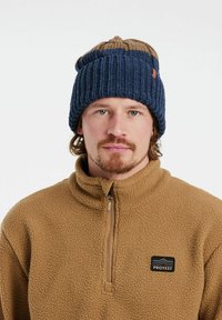 Blaue und braune gestrickte Mütze mit Rippstruktur und umgeschlagenem Saum, getragen von einer Person in einem beigen Fleece-Pullover mit Logopatch.