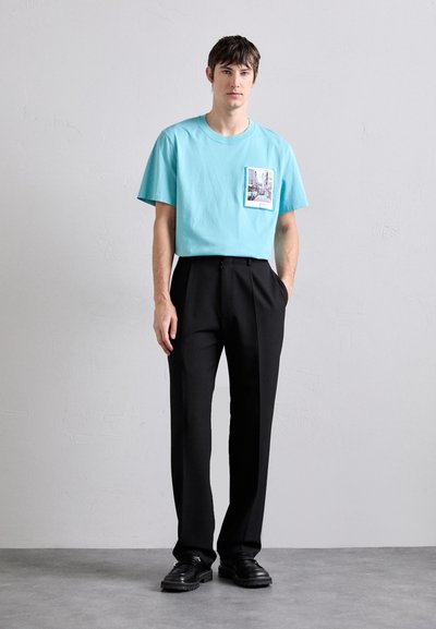 Tirkizna pamučna majica s džepom na prsima koji sadrži fotoprint, u kombinaciji s crnim plisiranim hlačama i crnim cipelama.