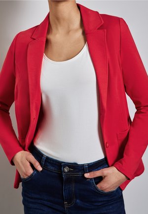 Osoba nosząca czerwony blezer na białym topie, z rękami w kieszeniach ciemnoniebieskich jeansów na neutralnym tle.